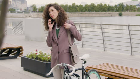 curly woman in formal clothes and bicyle talking on mobile phone on the city bridge