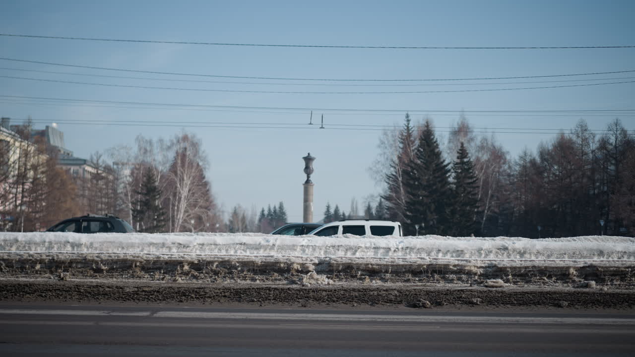 sunny winter city scene with van passing along two way road while cars move on opposite side, snowbanks line street beneath power wires, distant buildings and trees frame commute under clear blue sky