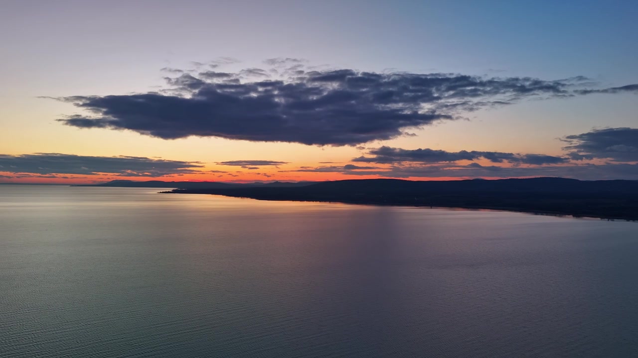 Eine Vogelperspektive über dem Plattensee in Ungarn fängt die ruhige Süßwasserfläche entlang der Nordküste in der Dämmerung ein, wobei farbenfrohe Himmelsreflexionen mit den fernen Hügeln und der Küstenlinie verschmelzen.