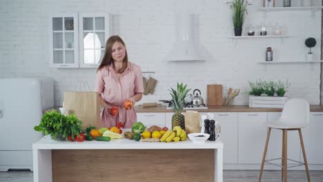 Attractive woman taking out fresh apples from brown paper bag. Table full of natural fruit and vegetables. Young female cares about healthy nutrition at home.