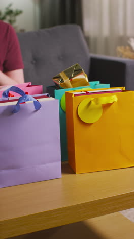 vertical video of man taking out gift wrapped presents from colourful bags with tag on table in lounge at home