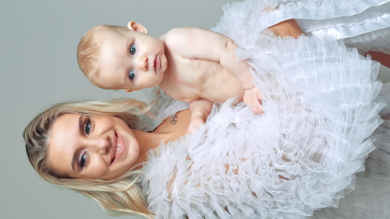 Mom and child portrait. Blonde woman holds her tiny grey-eyed boy smiling to camera. Vertical screen.