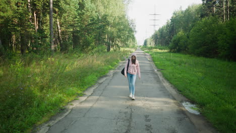 young woman in pink hoodie and jeans walking along empty forest road, holding her hoodie rope thoughtfully, casting long shadow on pavement under morning sunlight, surrounded by trees and wild grass