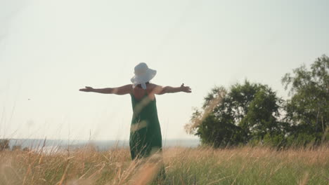 woman in green dress wearing wide hat spreads arms joyfully dancing in tall golden grass under warm sunlight, expressing freedom and happiness on peaceful adventure in tranquil open countryside