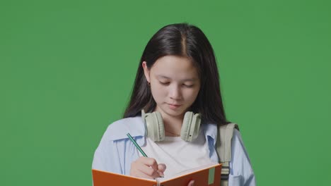 close up of asian teen girl student with a backpack smiling and taking note on notebook while standing in the green screen background studio