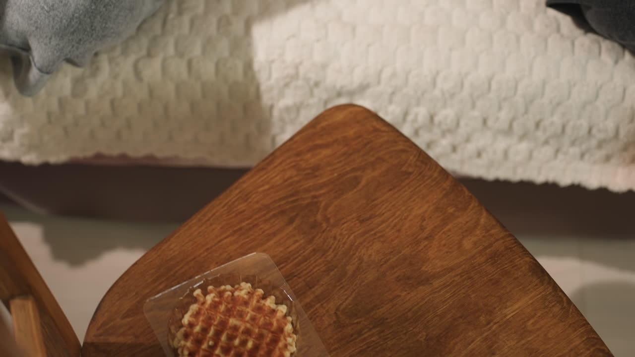 Unas manos blancas pasan una taza de té color ámbar sobre una mesa de madera con un gofre empaquetado, un desayuno informal, un gesto amable entre anfitrión e invitado, luz natural cálida, primer plano de un momento de hospitalidad en la mesa