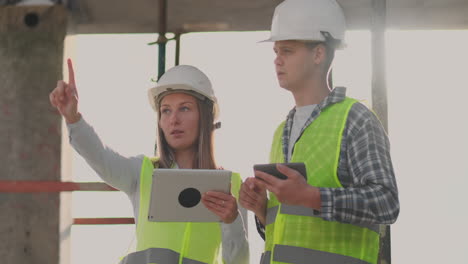 Ingenieros-Diseñadores-Un-Hombre-Y-Una-Mujer-Parados-En-El-Techo-De-Un-Edificio-En-Construcción-Y-Discuten-El-Plan-Y-El-Progreso-De-La-Construcción-Usando-Una-Tableta-Y-Un-Teléfono-Móvil.-Los-Constructores-Modernos-Discuten-La-Infraestructura-Del-Edificio-Y-Sus-Alrededores.