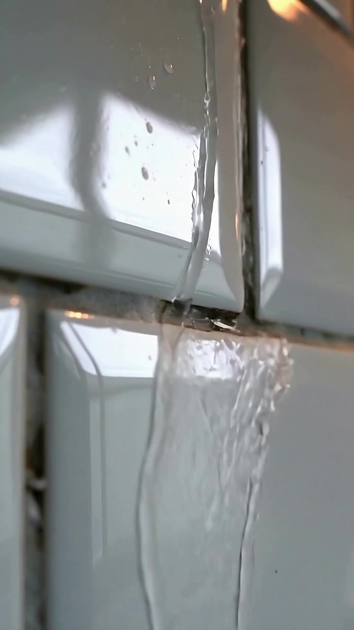 Vertical video: Flowing thin stream leaking from grout seam bulging into drops on kitchen tiles