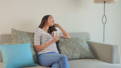 A-young-Asian-woman-sits-on-a-large-overstuffed-couch-holds-a-coffee-cup-sipping-it-and-smiling-every-once-in-a-while