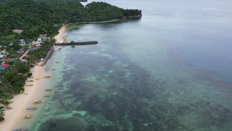 vista aérea ascendente de aguas prístinas del océano frente a botes bangka alineados en una idílica playa de arena blanca conectada a una comunidad rural en catanduanes, filipinas