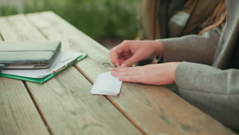 close side view of lady folding paper into plane while seated at wooden table outdoors, hands carefully shaping paper edges with documents and notebook placed nearby in natural light
