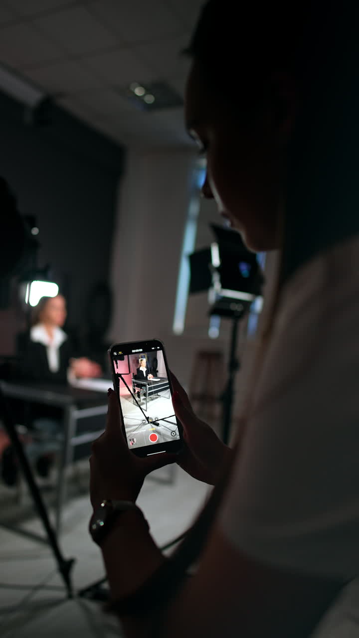 Brunette girl stands bent holding a phone and taking video. Unrecognized woman in black jacket sits at blurred backdrop talking. Vertical video.