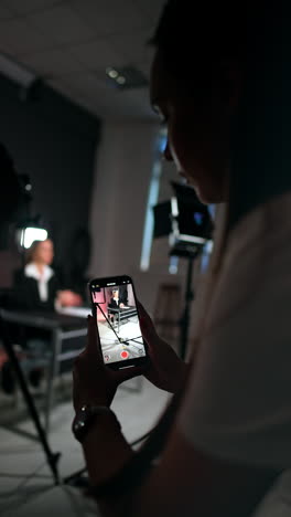 brunette girl stands bent holding a phone and taking video. unrecognized woman in black jacket sits at blurred backdrop talking. vertical video.