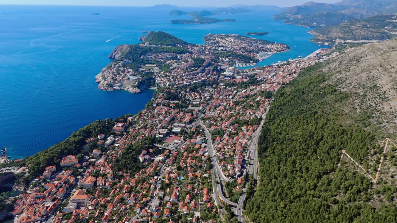 Aerial wide-angle landscape drone pans left from Mount Srđ over Dubrovnik, capturing terracotta rooftops, the deep blue Adriatic Sea, and rugged coastline stretching along the scenic Mediterranean
