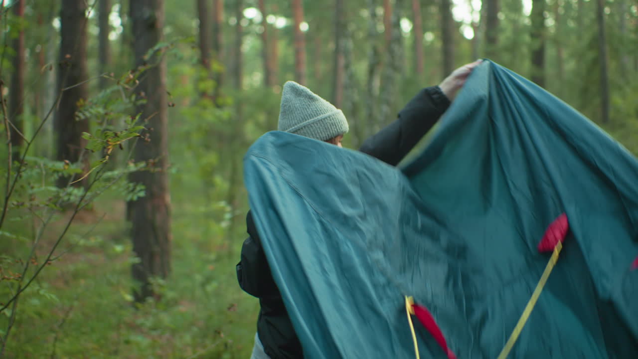 Young woman in black jacket and gray beanie holds tent cover in hand, smiling toward companion as she prepares to drape it over tent in peaceful forest with soft sunlight filtering through trees