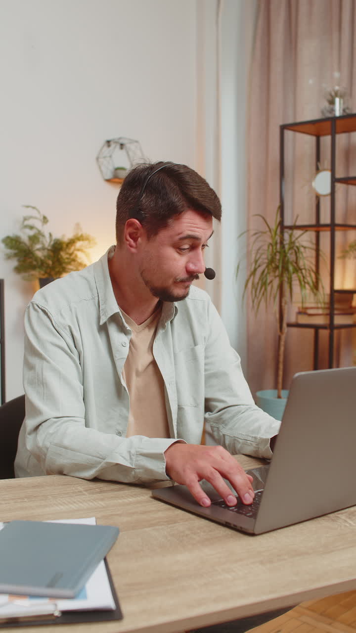 Young male customer service representative with headset using laptop computer at home office table