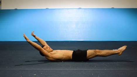 a guy doing an abs workout in a gymnastics gym doing lying down shortless leg and hand raises still shot