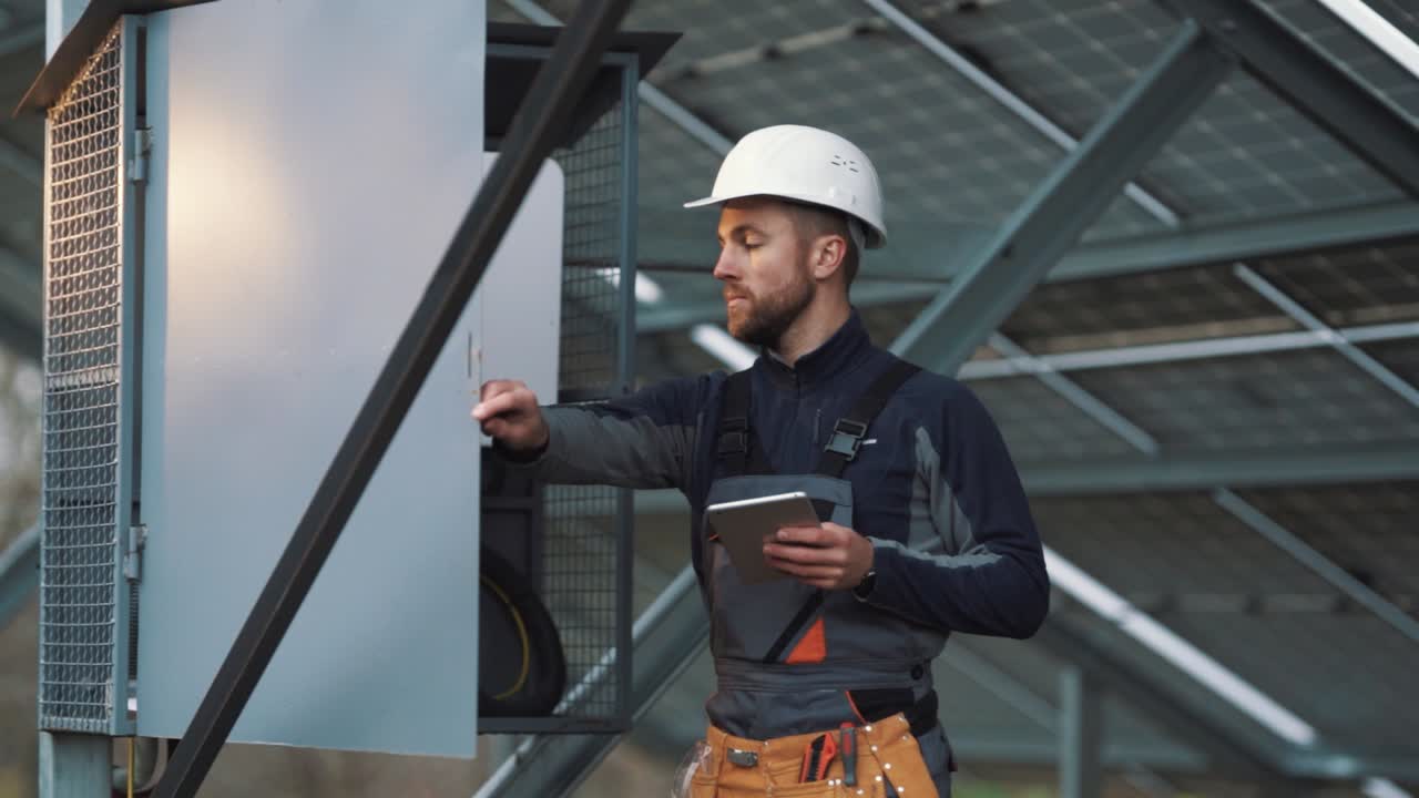 A professional man in a special electrician's uniform opens the switchboard and conducts a technical inspection with a tablet