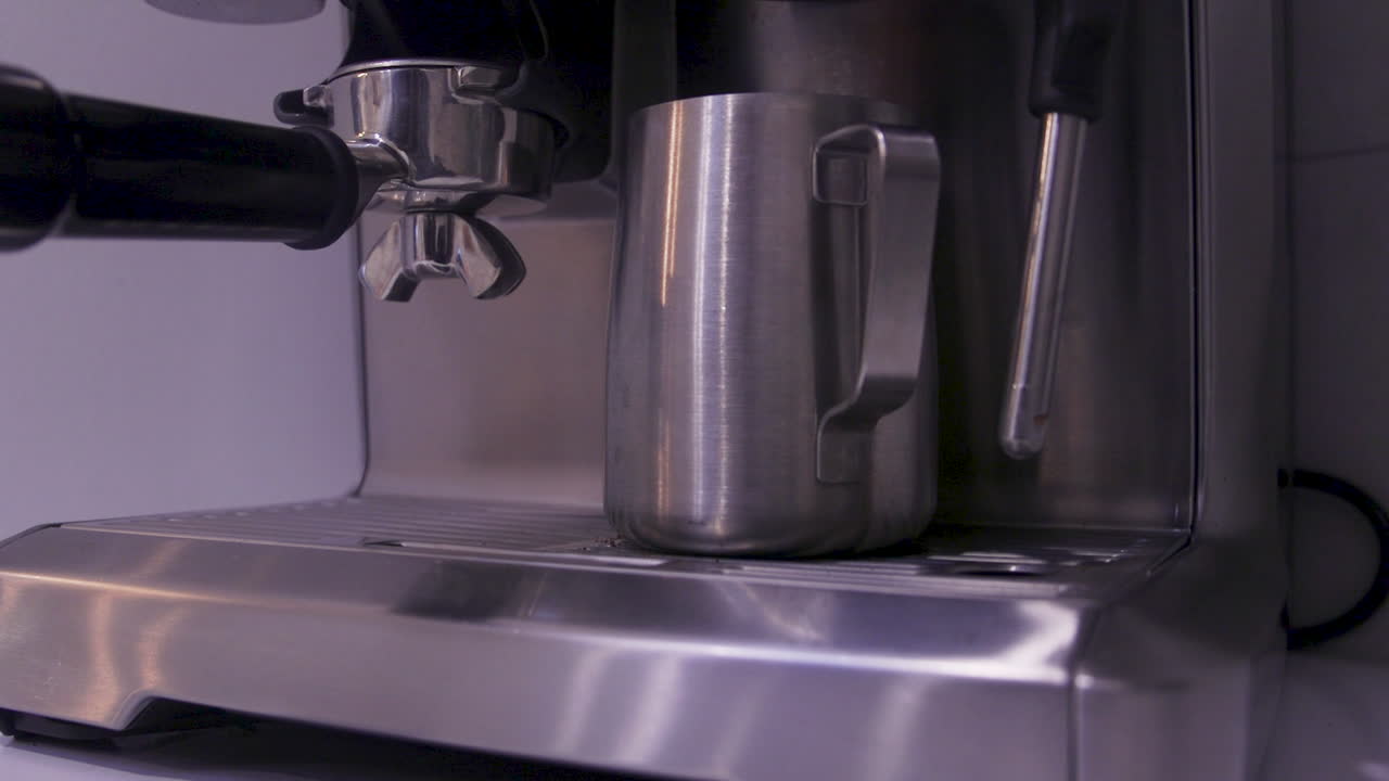 A hand removes a milk frothing jug from a coffee machine in a kitchen. The process shows maintenance of equipment used for preparing coffee drinks. Clear focus on the jug and machine parts