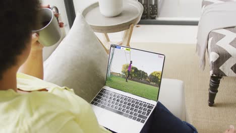 Video-of-biracial-woman-sitting-on-sofa-and-watching-football-on-laptop-at-home