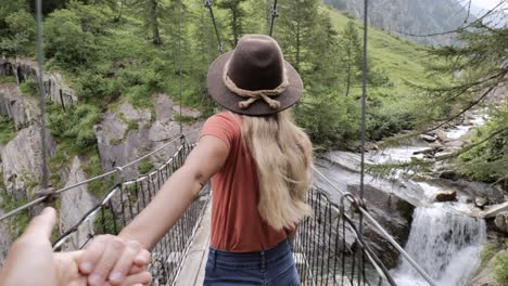 follow me to, woman holding companion's hand, leading him on a hike over suspension bridge in the forest