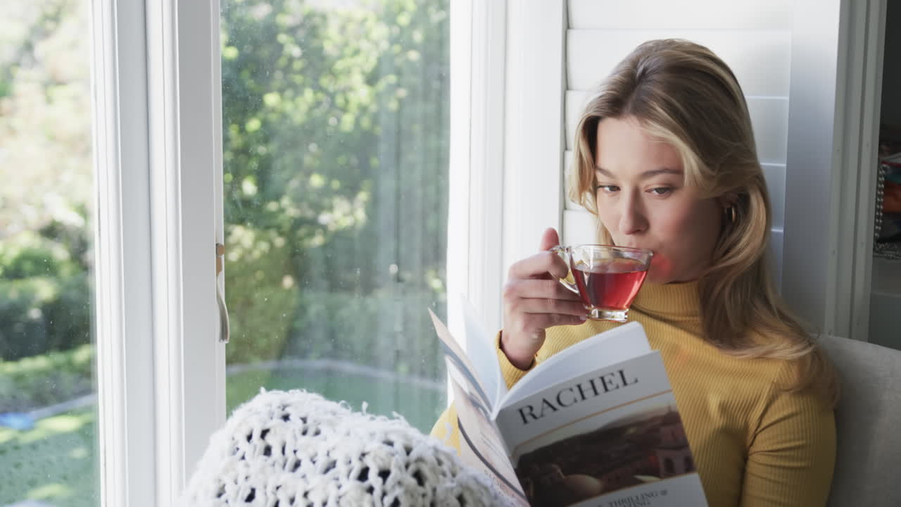 feliz mujer biracial sentada junto a la ventana bebiendo té y leyendo un libro en casa, en cámara lenta
