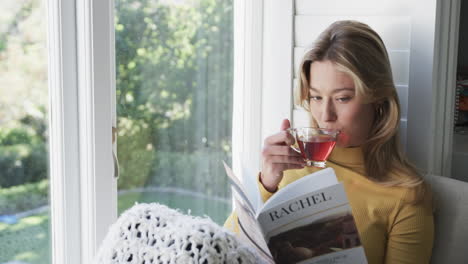 Feliz-Mujer-Birracial-Sentada-Junto-A-La-Ventana-Bebiendo-Té-Y-Leyendo-Un-Libro-En-Casa,-Cámara-Lenta