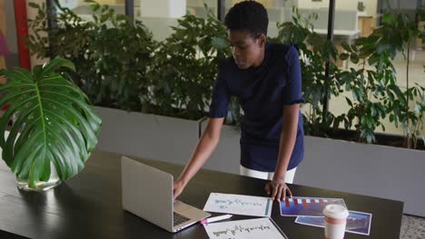 mujer de negocios afroamericana de pie usando una computadora portátil pasando por el papeleo en una oficina moderna