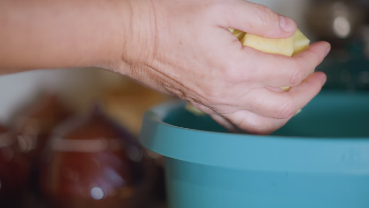 Escena de cocina con movimientos de manos firmes. Preparando ingredientes para guiso en una acogedora encimera de cocina. Primer plano de manos colocando cuidadosamente patatas en cubos en un cuenco turquesa bajo una cálida luz natural.