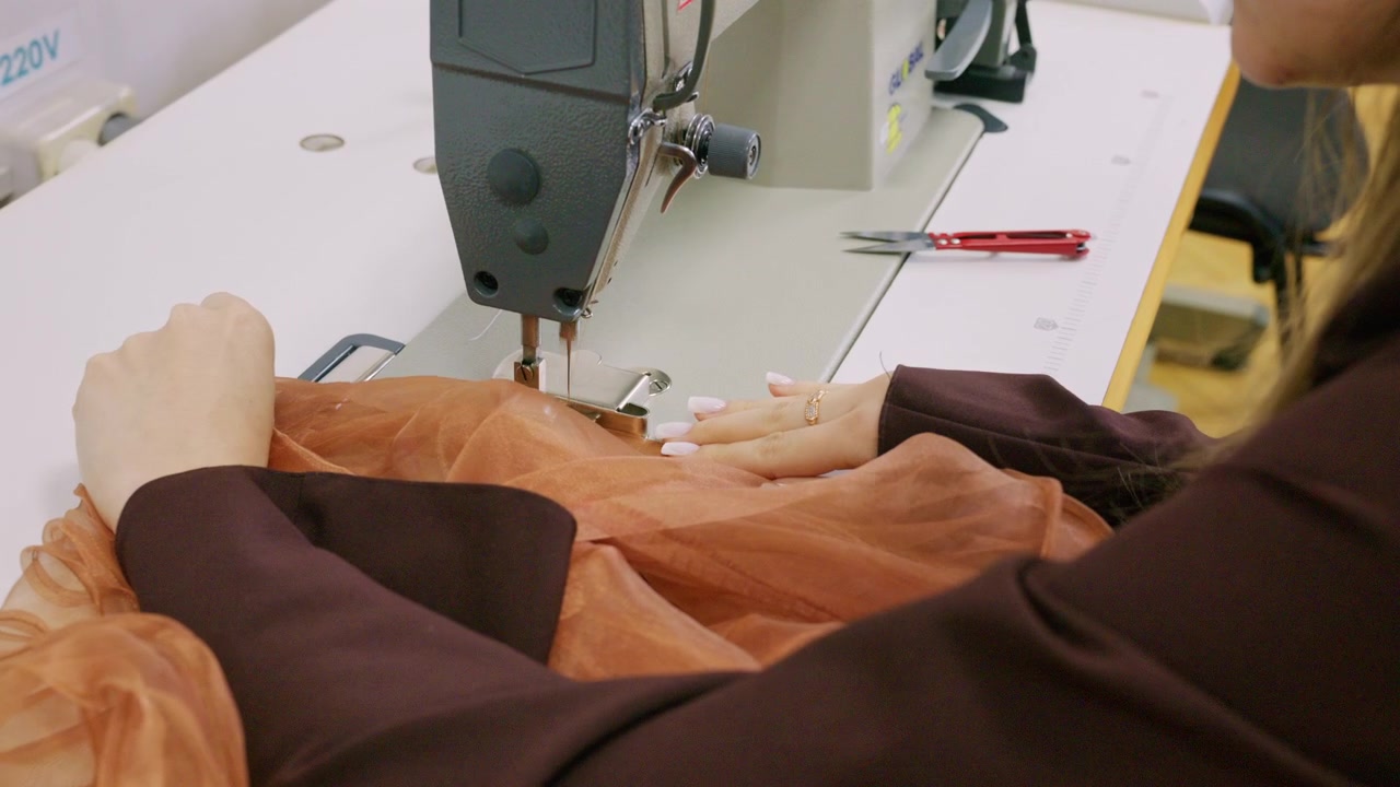 A fashion designer guides translucent orange organza fabric under the needle of an industrial sewing machine