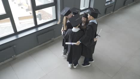 group of happy multiracial preschool students in mortarboard and gown. they are hugging, jumping and holding their diplomas.