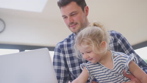 Padre-Trabajador-Usando-Una-Computadora-Portátil-En-Casa-En-El-Mostrador-De-La-Cocina-Mientras-Cuida-A-Su-Hija-Pequeña---Filmado-En-Cámara-Lenta