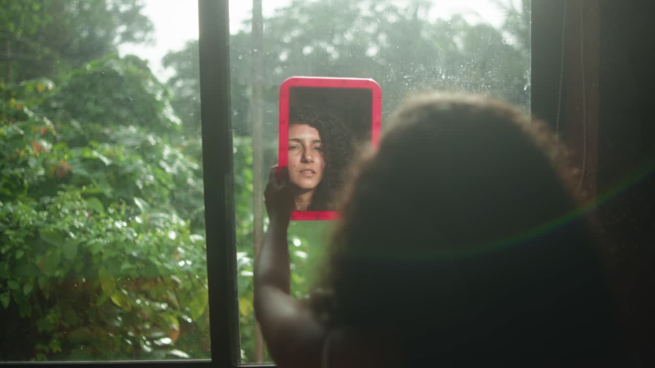 Artistic shot of a woman facing an open window, her face reflected in a mirror, surrounded by lush greenery