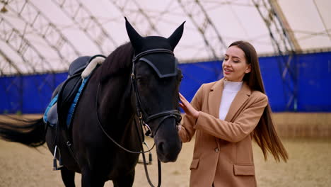 beautiful brunette woman in beige jacket holding a beautiful black horse. lady leading a graceful animal by the drill-hall. blurred backdrop.