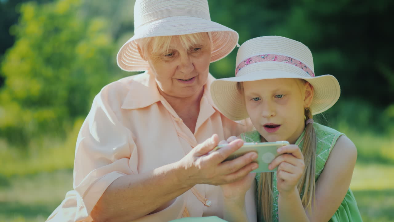할머니와 손녀는 스마트폰을 함께 사용하여 여름날 4k에 공원에서 휴식을 취합니다.