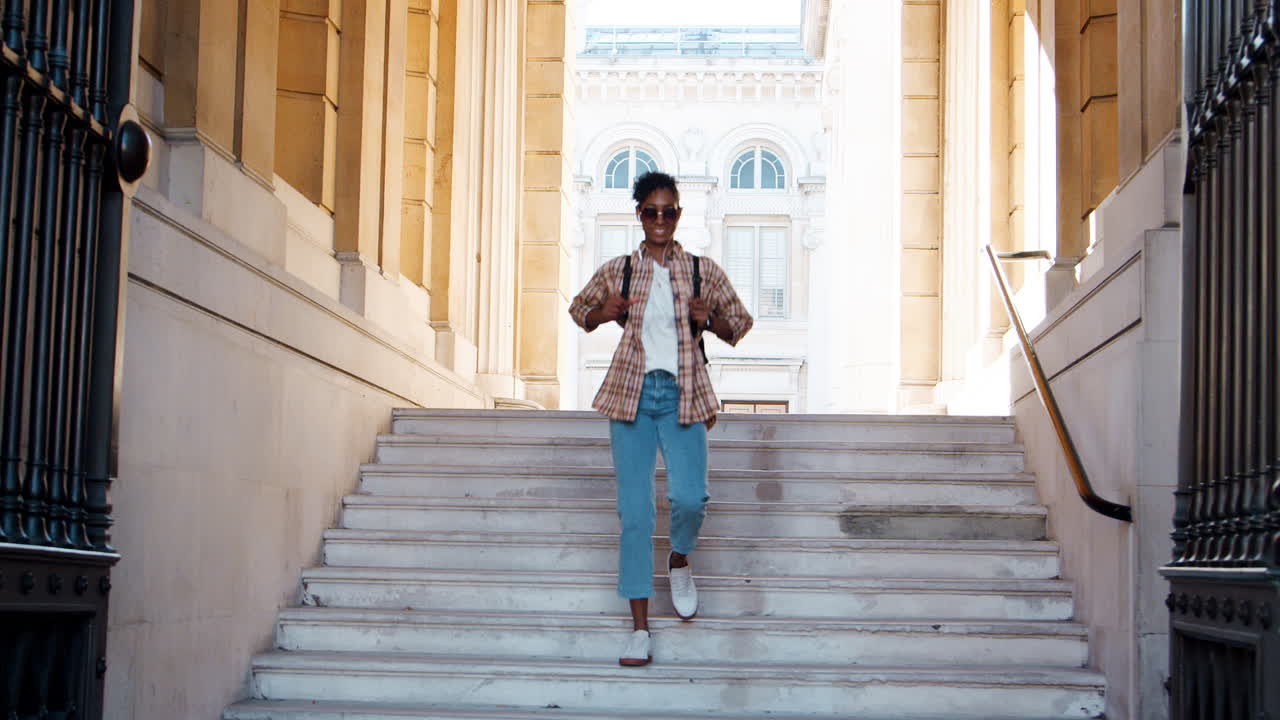Fashionable young adult woman wearing a plaid shirt  and blue jeans walking down steps outside a historical building complex listening to music with earphones, low angle