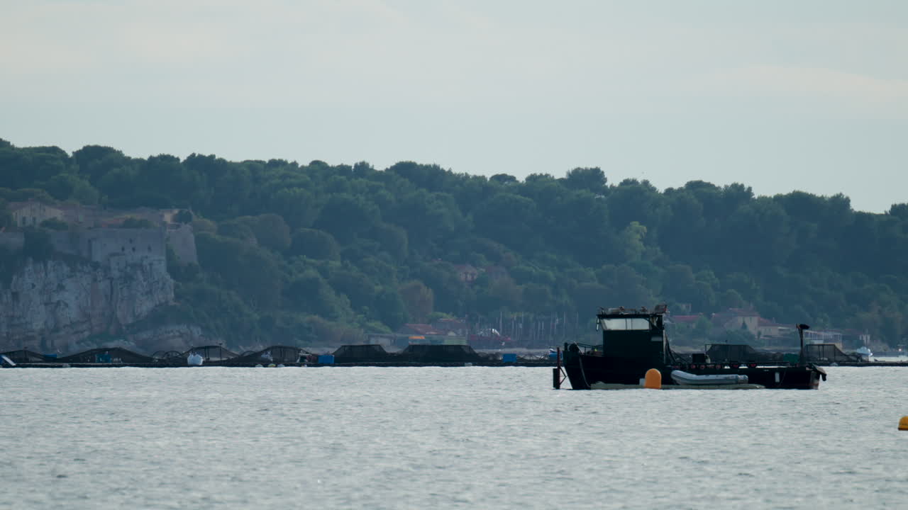 A black industrial boat floating beside offshore fish farming nets with forested hills in the background