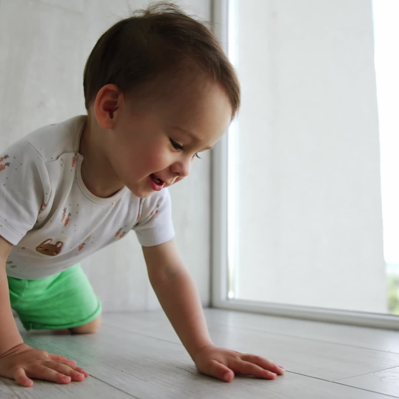 Adorable baby boy plays at home. Kid sits on the floor, puts his cheek to the floor and stands up again