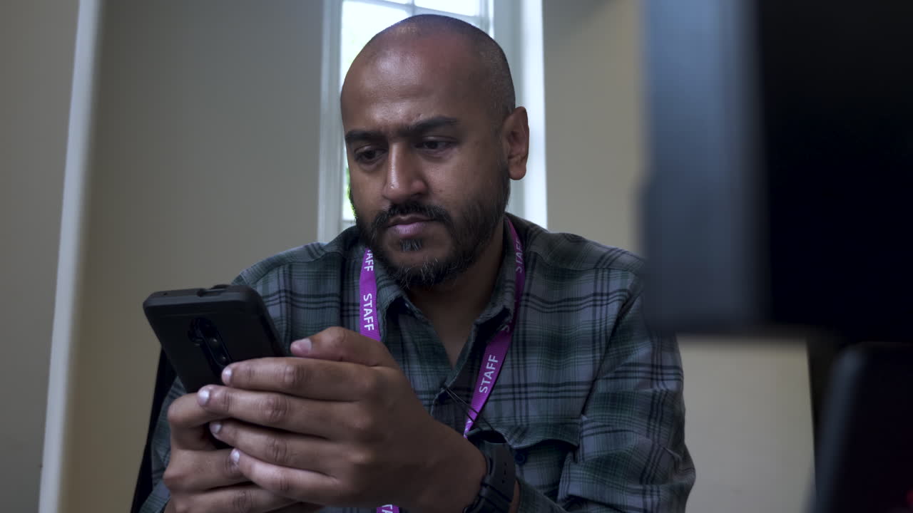 un primer plano desde detrás de una computadora de oficina mientras un indio asiático se sienta en su escritorio respondiendo a un mensaje de texto en su teléfono móvil