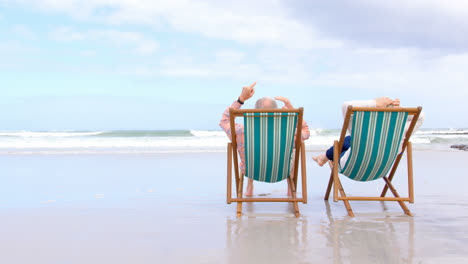 rear view of old caucasian senior couple sitting on sun lounger and holding hands at beach 4krear vi