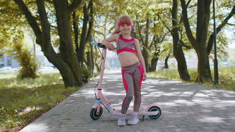 athletic fit young girl rides on electric scooter, standing near bike looking at camera in park