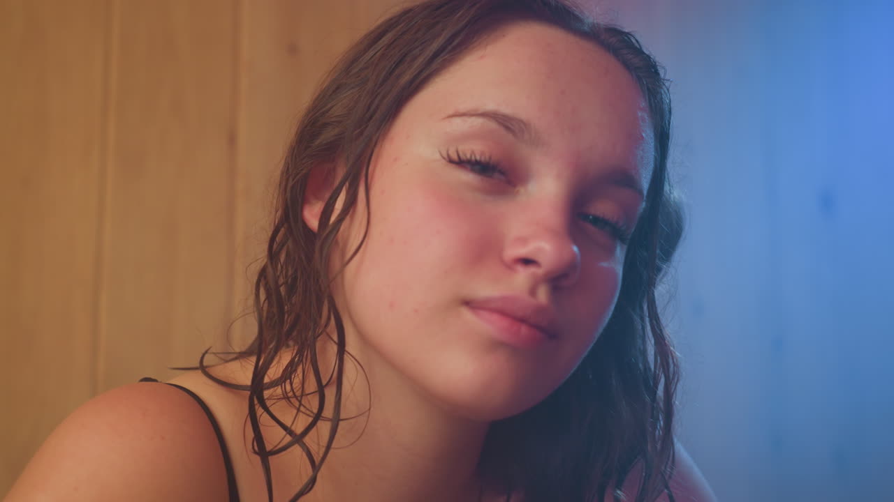 young woman wrapped in towel leans against wooden sauna wall, gazing with relaxed seductive expression, damp hair framing face under warm amber light, conveying intimate comfort