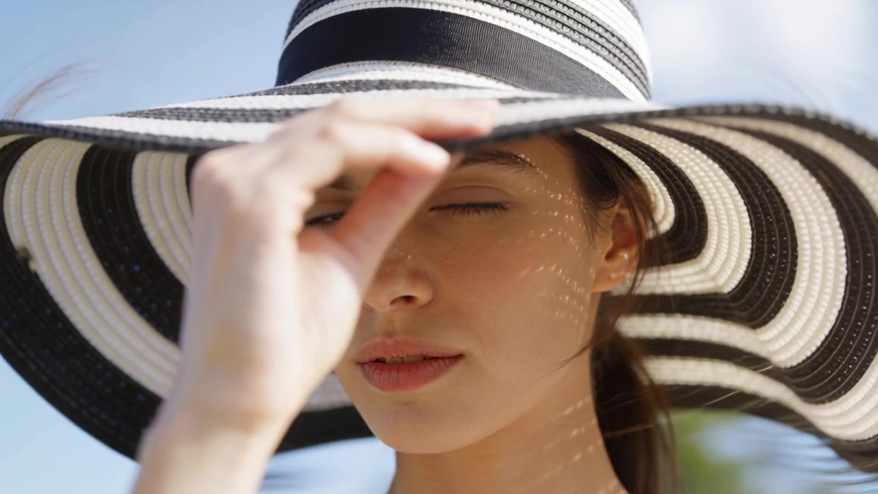 Donna che abbassa con la mano un cappello a tesa larga a righe, riparandosi il viso dal sole splendente all'aperto, facendo dei puntini.