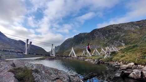 Woman-walking-on-small-wooden-suspension-bridge-while-crossing-small-mountain-lake---Hiking-trip-to-Kiellandbu-in-beautiful-mountain-landscape-Norway