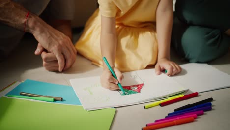 Close-up-shot-of-a-little-girl-in-a-yellow-dress-drawing-a-house-using-multi-colored-markers-on-a-piece-of-white-paper-while-sitting-on-the-floor-with-her-parents-in-a-modern-room