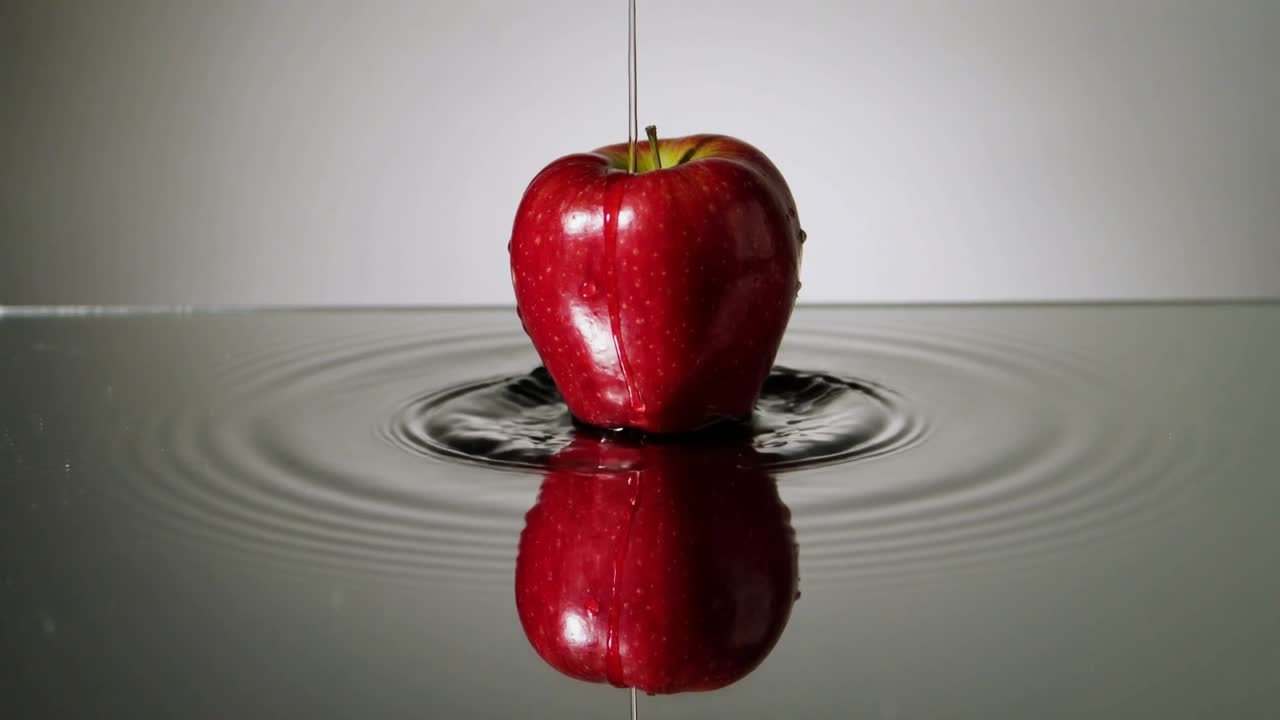 Dryppende topstrøm rammer skinnende rødt æble på studiebassin, skaber ringe i vandet og spejlsløring.