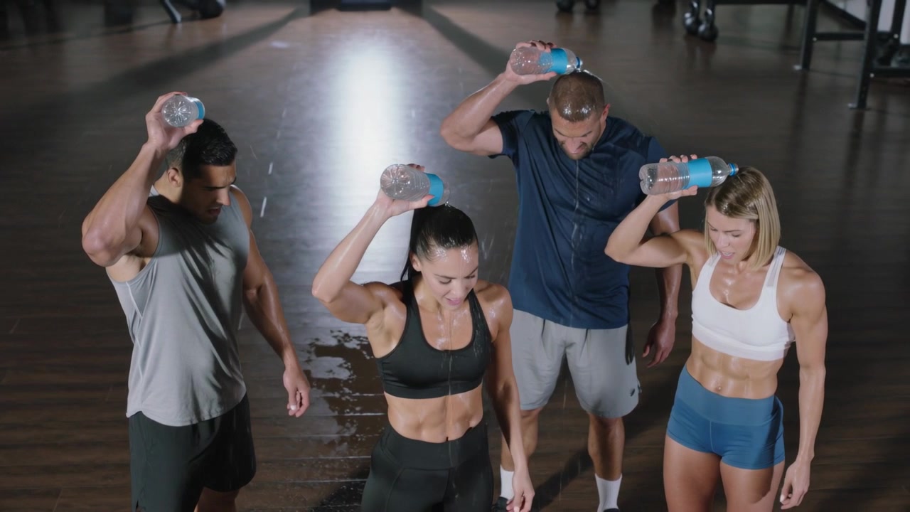Cuatro atletas volcando botellas sobre sus cabezas a la señal durante un entrenamiento en el gimnasio, mojando la ropa deportiva y el suelo.