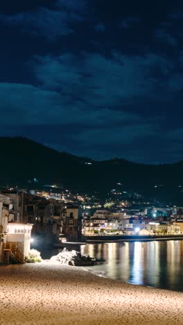 vertical video of beach timelapse at night. cefalu city lights by the coast. clouds moving. people walking. reflections in the water.