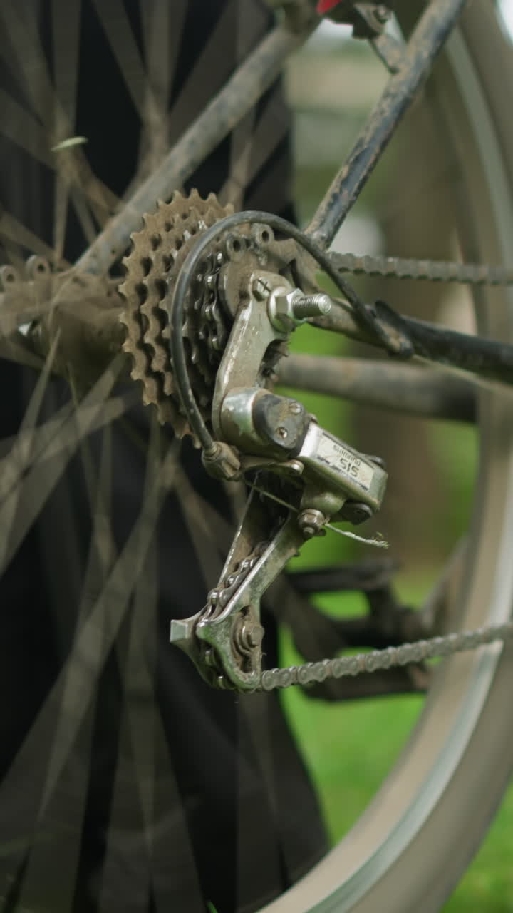 primer plano de un individuo con pantalones y zapatillas negras de pie al lado de una bicicleta estacionada en un campo cubierto de hierba, ajustando el pedal hacia atrás, levantando el neumático trasero y pedalando para girar la rueda