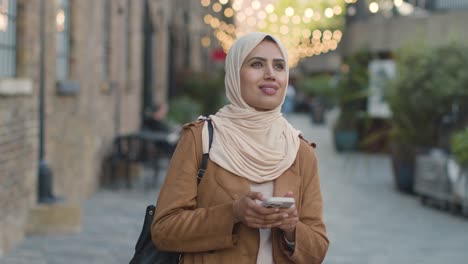 muslim woman wearing hijab walking along city street sending text message to date on mobile phone